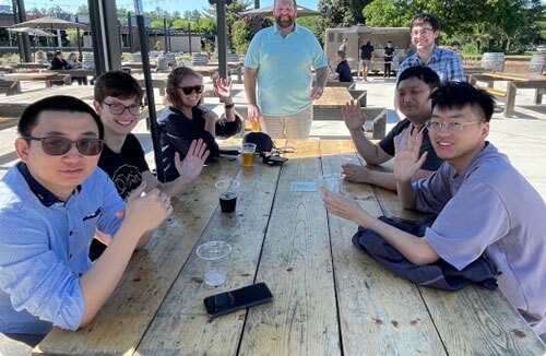 A group of people around a shaded picnic table, smiling and waving at the camera